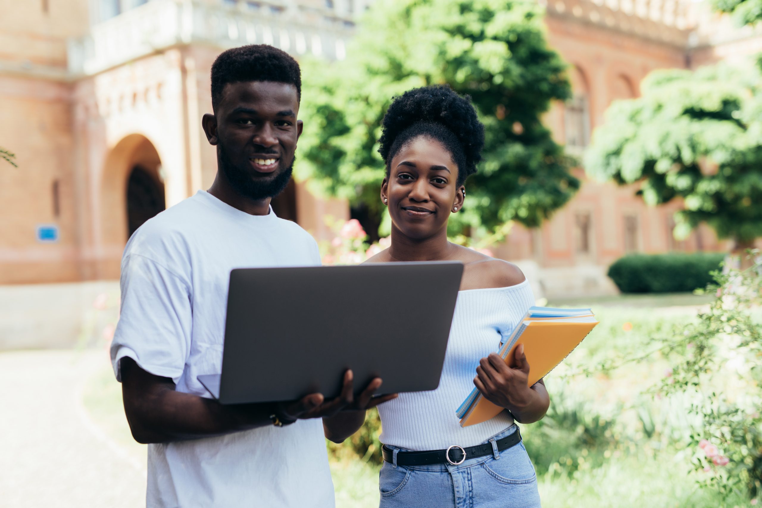 Two young students with laptop standing in campus yard