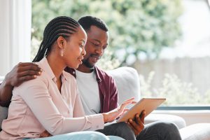 Couple noir souriant assis sur un canapé à la maison, avec tablette, naviguant sur Internet pour trouver des options d'assurance habitation. Famille, relations et satisfaction sur le site web pour une assurance vie et une couverture obsèques.