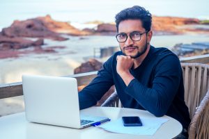 Bel homme indien prospère, élégant et bien habillé, travailleur indépendant travaillant avec un ordinateur portable sur la plage. Travail indépendant et à distance. Étudiant en affaires dans un café d'été au bord de l'océan indien