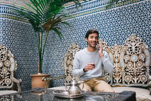 Pleased ethnic male tourist in casual clothing calling on smartphone at exotic cafe