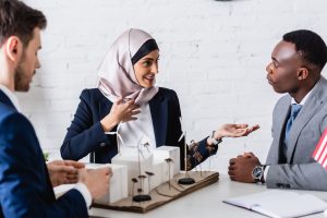 Une femme d'affaires arabe souriante montre du doigt un partenaire commercial afro-américain près d'un modèle de centrale électrique verte, premier plan flou