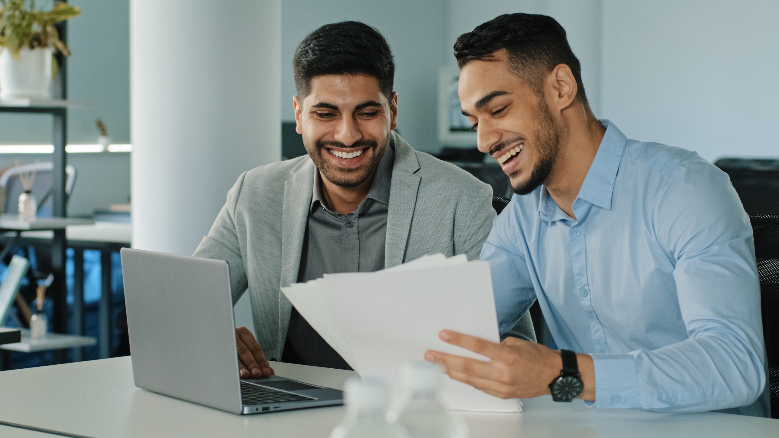 Smiling young colleagues holding paper documents, Indian employees sitting in office with laptop receiving good news. Two successful Arab male businessmen satisfied excellent result of negotiations