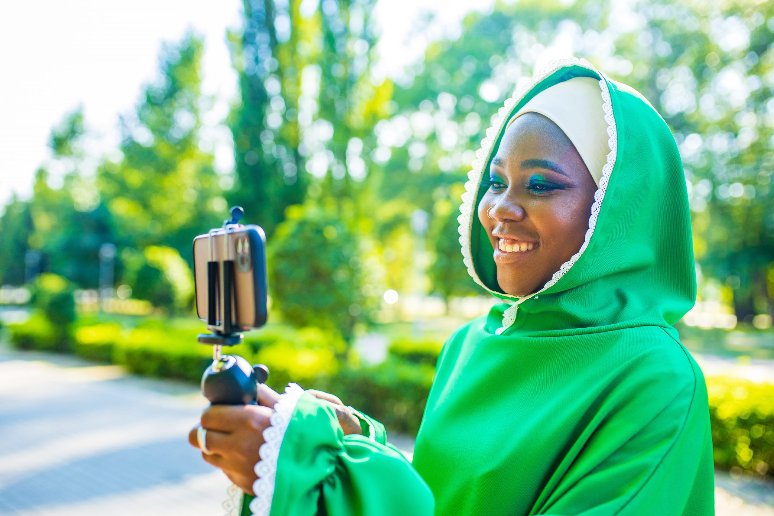 latin hispanic wonan in green muslim hijab with bright make up and nose piercing taking video stream outdoors summer park.