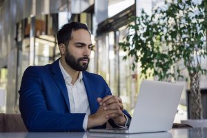 Bel homme d'affaires marocain utilisant un ordinateur portable pour suivre des formations en ligne dans un bureau moderne. Jeune cadre moyen-oriental pensif, planifiant un projet, assis sur son lieu de travail.