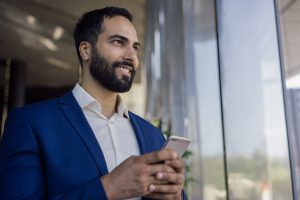 Bel homme d'affaires marocain, smartphone à la main, faisant des achats en ligne, regardant la vitrine, debout dans un bureau moderne. Homme du Moyen-Orient utilisant une application mobile, espace de copie. Concept d'entreprise réussi.