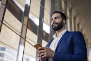 Bel homme d'affaires moyen-oriental souriant utilisant son téléphone portable dans un bureau moderne. Responsable arabe confiant, en tenue de soirée, en train d'envoyer des SMS par la fenêtre. Entreprise prospère.