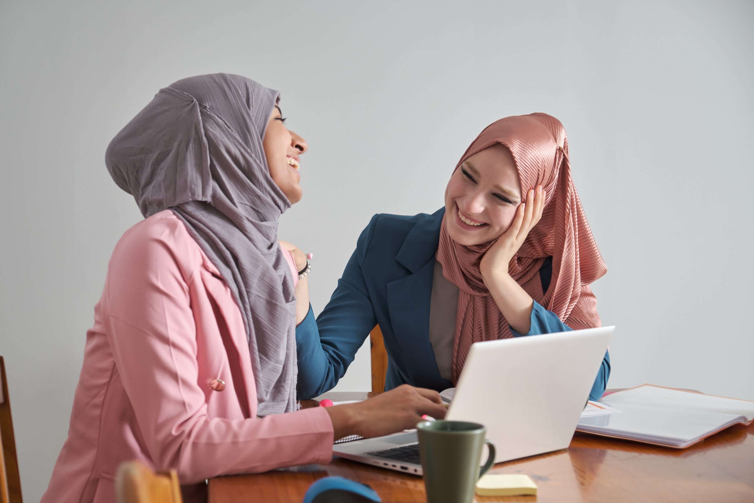 Two muslim business women wearing hijab laughing and working in a laptop.