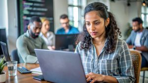 Un groupe de personnes est assis à une table avec des ordinateurs portables dans un environnement collaboratif. Une personne au premier plan est concentrée sur son ordinateur, tandis que d'autres travaillent en arrière-plan. Généré par l'IA.